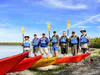 Group of men wearing life jackets stand together for a photo on a beach with kayaks visible in the foreground.